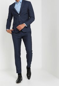 Abito navy con un sottile motivo a quadri, design aderente, giacca a un bottone e pantaloni tapered. Abbinato a una camicia azzurra chiara e scarpe nere.