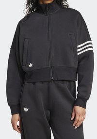 Donna che indossa una giacca a zip nera e pantaloni coordinati con i loghi trefoil bianchi di Adidas e tre strisce bianche su una manica.