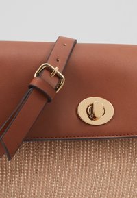 Borsa in pelle marrone con pattina, chiusura a girello in tonalità oro e tracolla regolabile, appoggiata su materiale beige intrecciato e testurizzato.