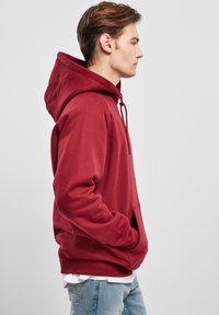 Hoodie bordeaux con cappuccio regolabile, tasca frontale, polsini a costine e texture morbida, indossato sopra una camicia bianca a strati e jeans blu.