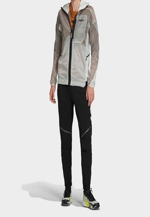 Person trägt eine graue leichte Kapuzenjacke, schwarze Sporthose mit reflektierenden Details und schwarz-gelbe Laufschuhe, steht.