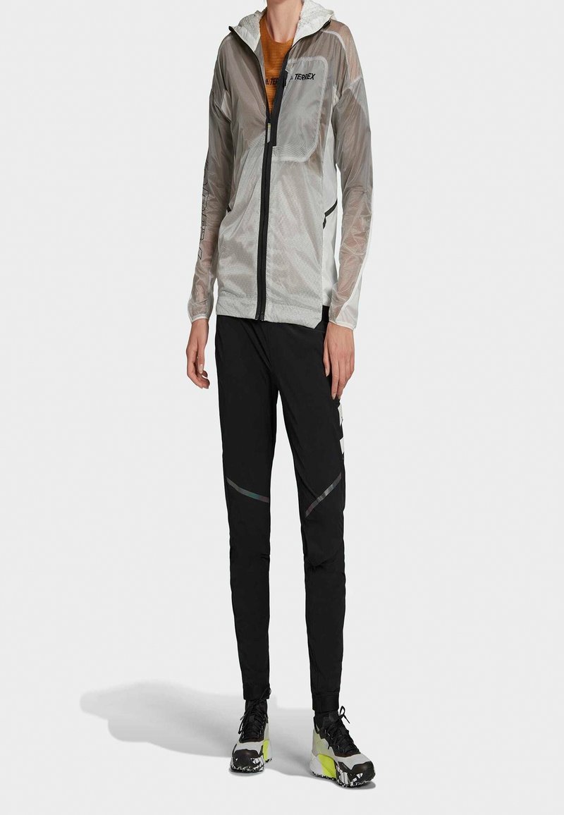Personne portant une veste légère à capuche grise, un pantalon de sport noir avec des détails réfléchissants et des chaussures de course noires et jaunes, debout.