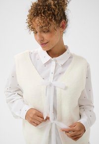 Biały, puszysty kamizelka z dekoltem w serek i przewiązaną z przodu kokardą, noszona na białej koszuli z małymi czarnymi literami nadrukowanymi.