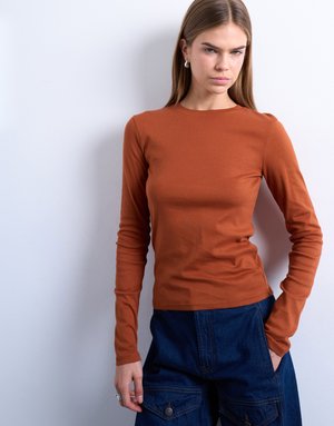 Vrouw met steil lichtbruin haar, gekleed in een roestkleurig shirt met lange mouwen en donkerblauwe broek, staand tegen een witte muur.