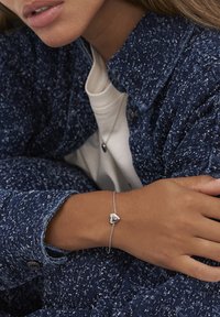 Person som bär en strukturerad blå jacka och vit skjorta med matchande silverfärgad hjärtformad armband och halsband, vilar armen över kroppen.