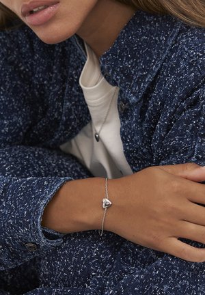Person som bär en strukturerad blå jacka och vit skjorta med matchande silverfärgad hjärtformad armband och halsband, vilar armen över kroppen.