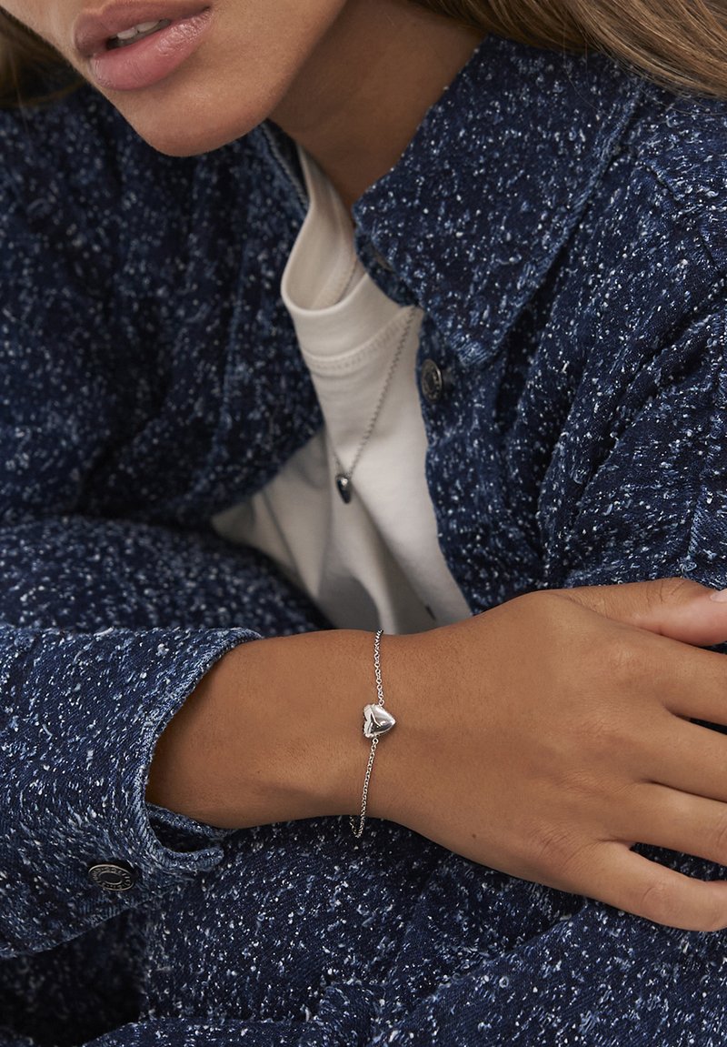 Person som bär en strukturerad blå jacka och vit skjorta med matchande silverfärgad hjärtformad armband och halsband, vilar armen över kroppen.