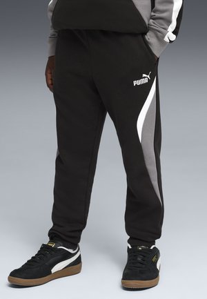 Person trägt schwarze Puma-Jogginghose mit grauen und weißen Seitenpaneelen sowie schwarze Puma-Sneaker mit weißen Streifen.