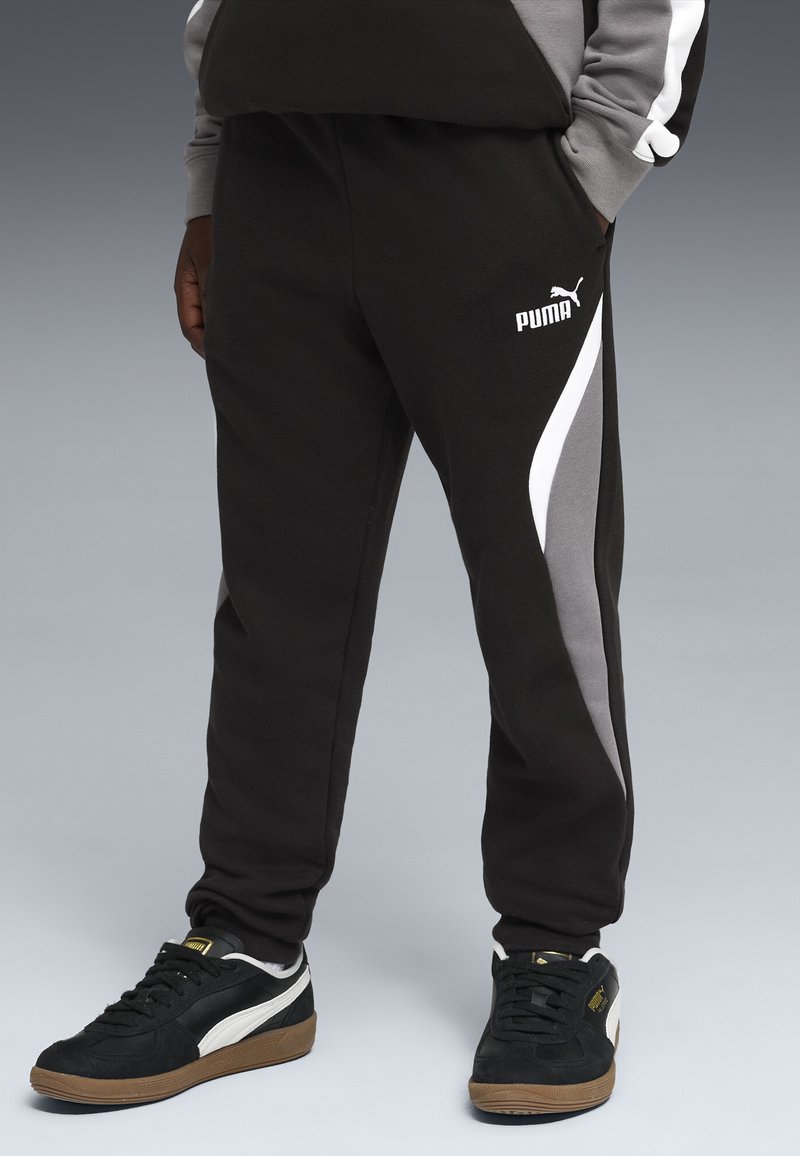 Persoon draagt zwarte Puma joggingbroek met grijze en witte zijpanelen en zwarte Puma sneakers met witte strepen.