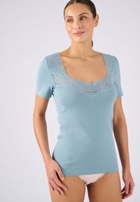 Camicetta azzurra a maniche corte realizzata in tessuto morbido, con inserto in pizzo e bordo ondulato attorno a un scollo quadrato.