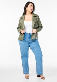 Olive green faux leather biker jacket, white fitted top, light blue textured straight-leg jeans, and brown block-heeled sandals.