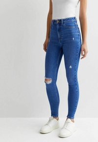 Jeans de mezclilla azul ajustados con una cintura alta y detalles desgastados en las rodillas. Combinados con zapatillas blancas. Textura suave y diseño ceñido.