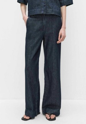 Henkilö, jolla on yllään tummat leveälahkeiset farkut, vastaavat napitettava paita ja mustat neliövarvassandaalit, seisoo valkoista taustaa vasten.