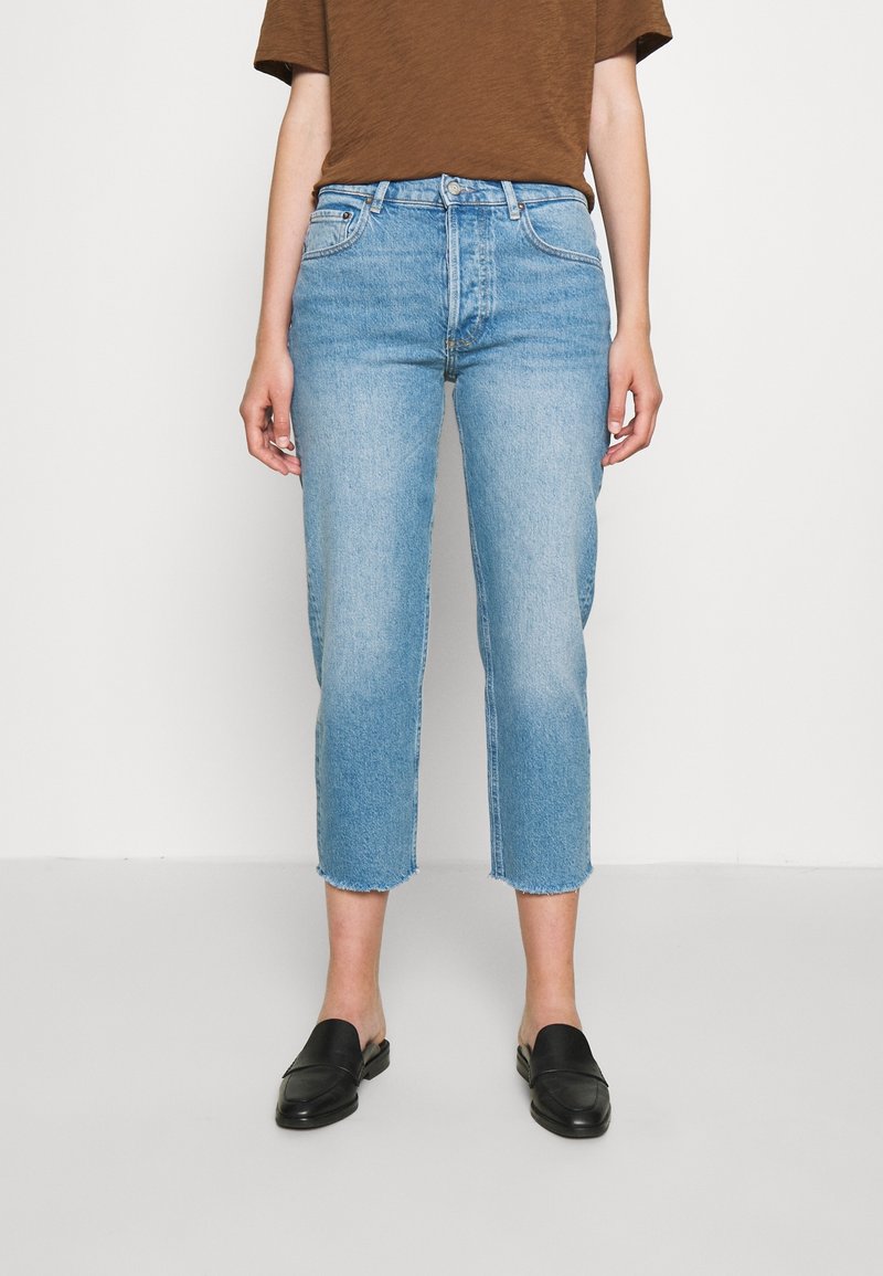 Vaqueros cortos de mezclilla azul claro con dobladillo sin rematar, que presentan un diseño estándar de cinco bolsillos, combinados con una camiseta marrón y mocasines negros.