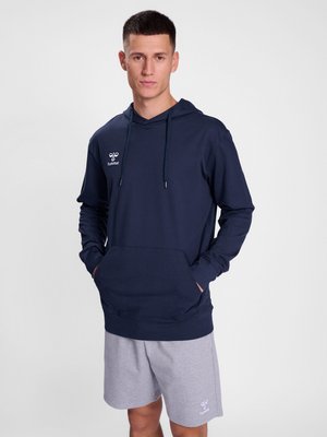 Jeune homme portant un sweat à capuche bleu marine et un short gris clair, debout les mains dans les poches du sweat à capuche, sur un fond uni.