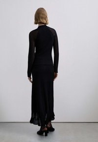 Mujer con cabello rubio corto que lleva un top negro de manga larga con textura acanalada y una falda a juego hasta el tobillo con detalles transparentes, de pie sobre un suelo gris.