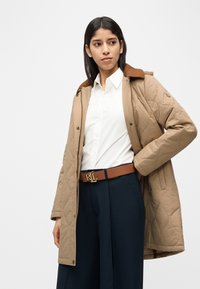 Καπιτονέ μπεζ σακάκι με καφέ γιακά, φορεμένο πάνω από λευκό πουκάμισο και σκουρόχρωμο παντελόνι. Διαθέτει καφέ ζώνη με ασημένια αγκράφα με λογότυπο.