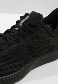 Chaussure de sport noire avec une tige en maille, surface texturée, lacets structurés et semelle rembourrée. Marque Nike en noir bien visible.