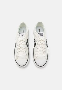 Weiße Leinenschuhe mit schwarzem Sternlogo und Akzenten, Gummi-Zehenkappe, Schnürverschluss und kontrastierendem schwarzen Rand um die Sohle.