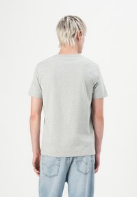 Persoană blondă cu părul scurt stând cu spatele, îmbrăcată cu un tricou simplu gri deschis și blugi din denim albastru deschis, pe un fundal alb.