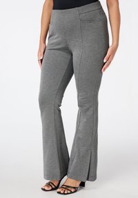 Femme portant un pantalon grisé évasé avec des poches latérales et des sandales à talons noires à lanières, se tenant devant un fond blanc.