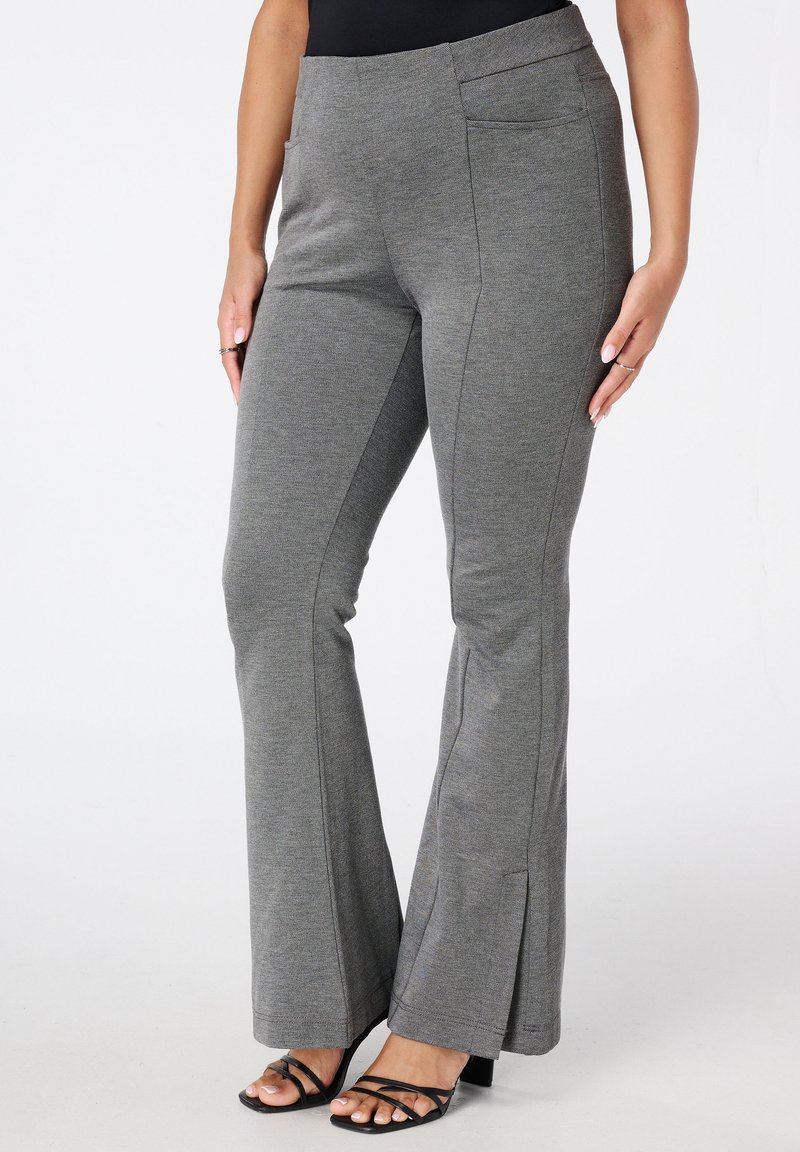 Femme portant un pantalon grisé évasé avec des poches latérales et des sandales à talons noires à lanières, se tenant devant un fond blanc.