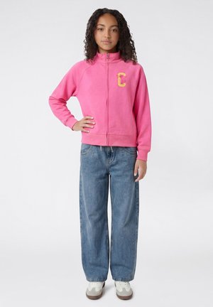 Junge Person mit lockigem Haar, trägt eine pinke Reißverschlussjacke, weite blaue Jeans und weiße Turnschuhe, steht mit einer Hand in der Hüfte.