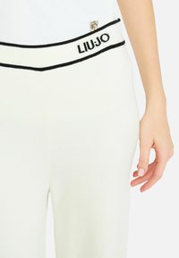 Bijele pletene hlače s crnim pojasom koji sadrži LIU JO logo. Glatka tekstura s prilagođenim oblikom. Mali ukrasni logo na pojasu.