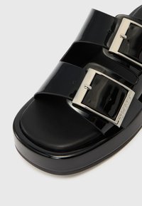 Sandalo a ciabatta in vernice nera con due ampi cinturini e fibbie argentate. Presenta una soletta con texture e una suola spessa e arrotondata.