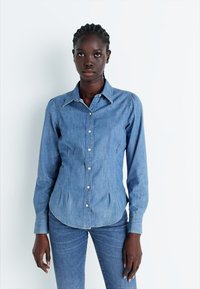 Jasnoniebieska koszula dżinsowa z punktowym kołnierzykiem, długimi rękawami i zapięciem na guziki, z subtelnymi pionowymi szwami i zaokrąglonym dołem.