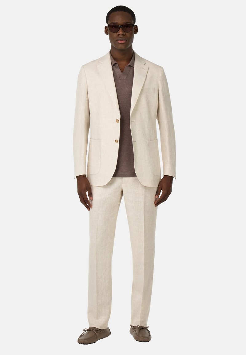 Traje de lino en beige claro con dos botones, combinado con una camiseta polo color marrón oscuro. Unos zapatos marrones sin cordones completan el atuendo.