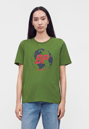 Jeune femme aux longs cheveux noirs porte un t-shirt vert avec un motif de globe et des lettres rouges "BP", associé à un jean bleu, sur un fond uni.