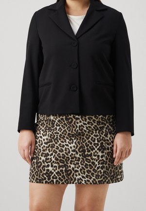 Personne portant un blazer noir boutonné sur un haut blanc et une jupe imprimé léopard, debout devant un fond uni.