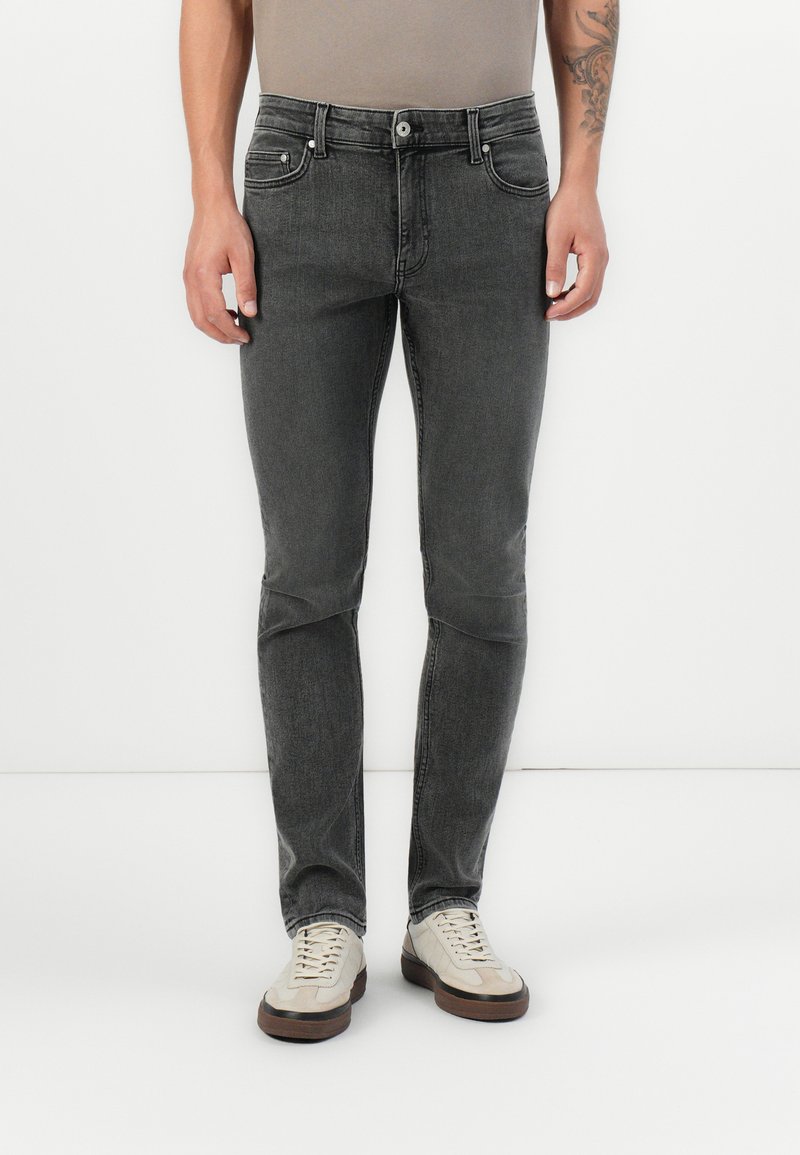 Szürke farmernadrág, slim fit fazonban, cipzáras nyitással, öt zsebbel, és diszkrét koptatással elöl. Világos sportcipővel viselve.