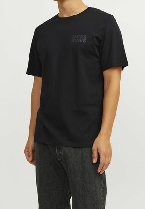 Man draagt een zwart Jack & Jones T-shirt met korte mouwen en donkergrijze jeans en staat tegen een effen lichte achtergrond.