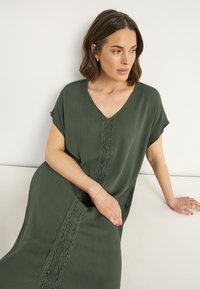 Olivengrünes Kleid mit V-Ausschnitt, kurzen Ärmeln und Spitzenverzierungen in der Mitte. Aus strukturiertem Stoff, fließendes Design.