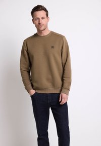 Felpa marrone lavorata a maglia con un motivo testurizzato, scollo rotondo e polsini a costine. Indossata con jeans blu scuro, caratterizzati da un piccolo dettaglio del logo.