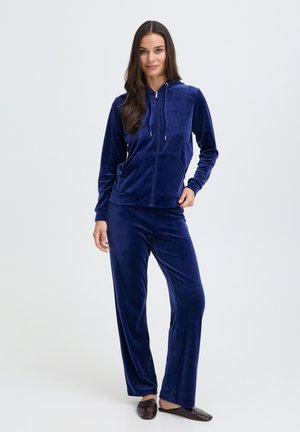 Femme portant un sweat à capuche en velours bleu foncé avec fermeture éclair et pantalon assorti, debout, détendue, une main dans la poche et un léger sourire.