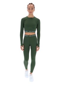 Žalias sportinis trumpas marškinėliai ir legginsai su prekės ženklo juostos dizainu. Modelis nešioja baltus sportbačius. Audinys atrodo lygus ir gerai prigludęs.