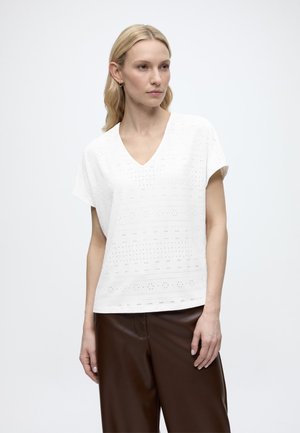 Blondynka w białej bluzce z krótkim rękawem i dekoltem w serek z ażurowymi wzorami oraz ciemnobrązowych spodniach, stojąca na jednolitym tle.