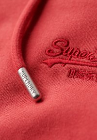 Rode hoodie met geborduurd "Superdry" logo. Heeft een gestructureerde afwerking en een zilveren metalen aglet aan de trekkoord.