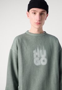 Zelená žebrovaná mikina s kulatým výstřihem a vybledlým bílým logem na hrudi. Nosí se s černou pletenou čepicí.