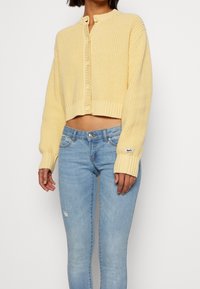 Gul, stickad croppad cardigan med rund halsringning, framknäppning, ribbade ärmslut och en etikett på ärmen, i kombination med ljusbl å jeans.
