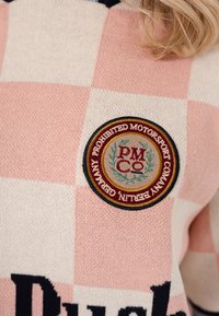 Pulover în carouri roz și crem, cu o insignă circulară brodată pe care scrie „Prohibited Motorsport Company Berlin Germany.”