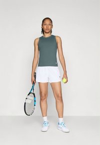 Top verde escuro sem mangas, calções brancos, sapatilhas de ténis azuis e brancas. Segura uma raquete de ténis e uma bola de ténis amarela.