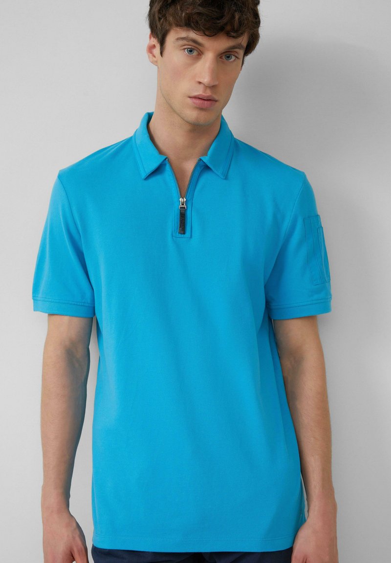 Turkusowa koszulka polo z krótkim zamkiem, krótkimi rękawami i kieszonką na lewym ramieniu. Gładka struktura, luźny krój.