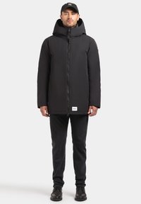 Juoda izoliuota parka striukė su aukštu apykakle ir užtrauktuku. Yra etiketė apačioje, dėvima su juodais džinsais ir batais.