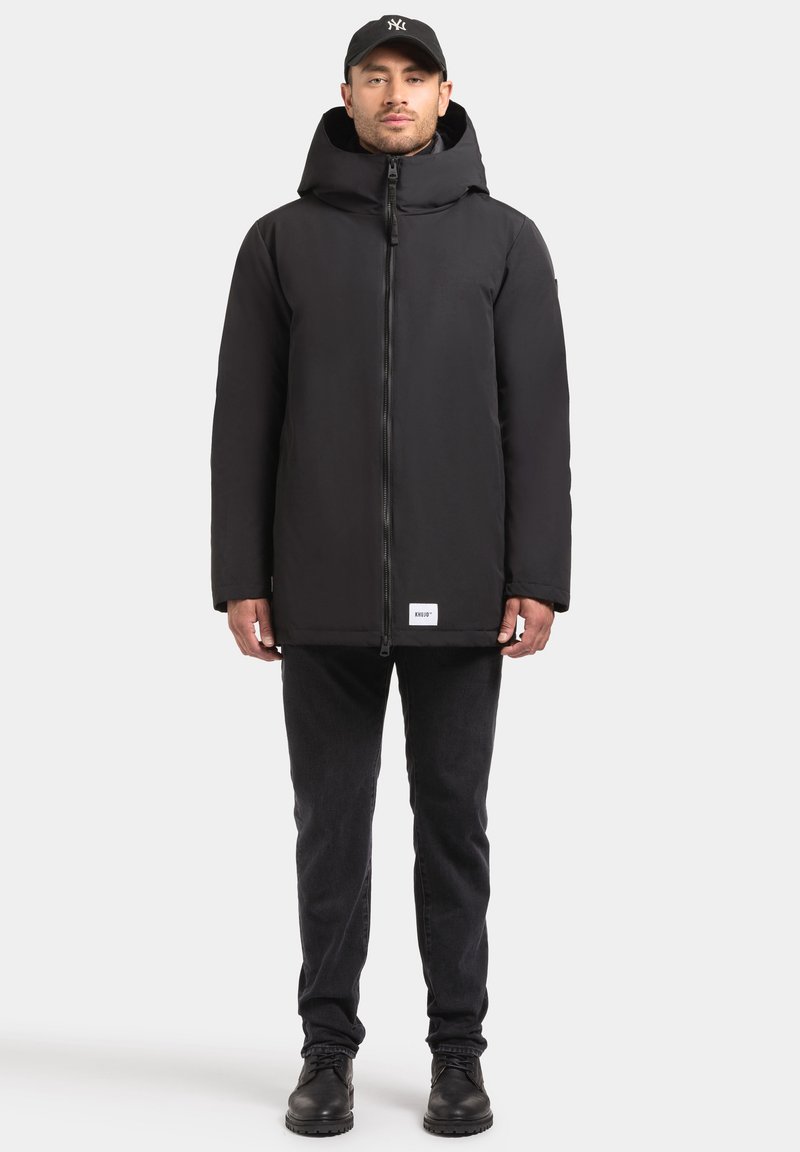 Juoda izoliuota parka striukė su aukštu apykakle ir užtrauktuku. Yra etiketė apačioje, dėvima su juodais džinsais ir batais.