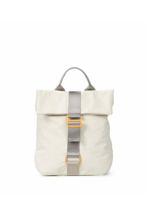 Persona con ropa informal sosteniendo una mochila color crema con correa gris y hebilla amarilla, vistiendo pantalones vaqueros holgados y sandalias.
