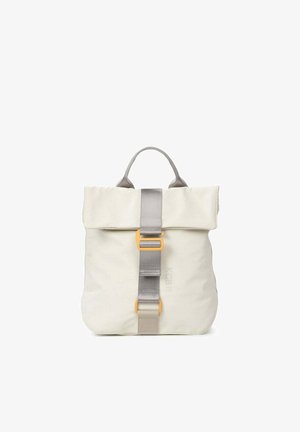 Person in casual outfit holding a cream-colored backpack with gray strap and yellow buckle, wearing loose denim pants and sandals.